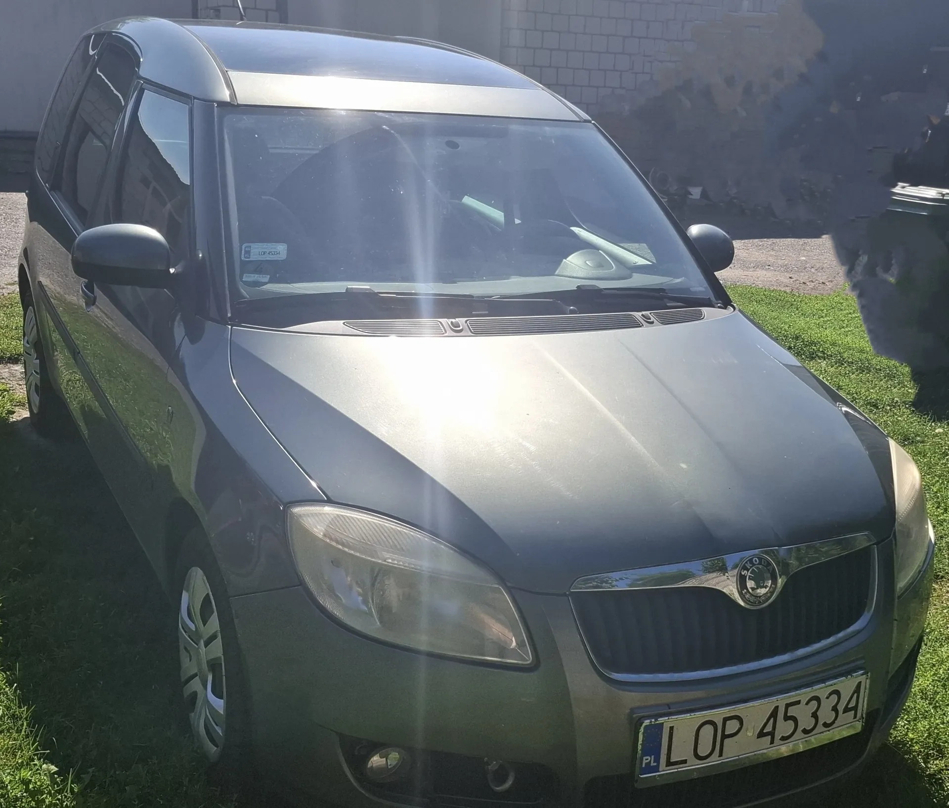 Szary samochód osobowy marki Skoda zaparkowany na trawie. Widoczny jest przód pojazdu z tablicą rejestracyjną o treści LOP 45334 oraz logo producenta na grillu.