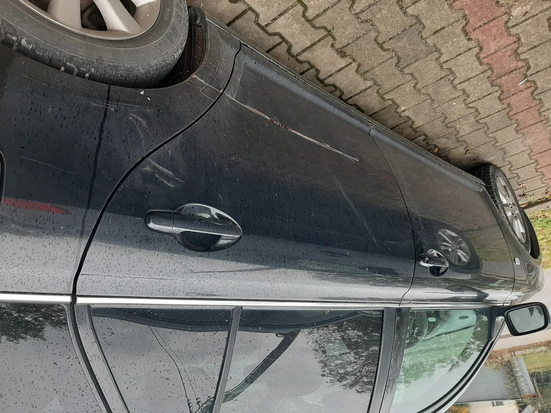 Ciemnoszary samochód osobowy zaparkowany na brukowanej powierzchni. Widoczne są zadrapania na bocznych drzwiach oraz krople deszczu na karoserii i szybach. W tle fragment ogrodzenia, drzewa i chmurne niebo. Samochód ma srebrne felgi i lusterko boczne po lewej stronie.