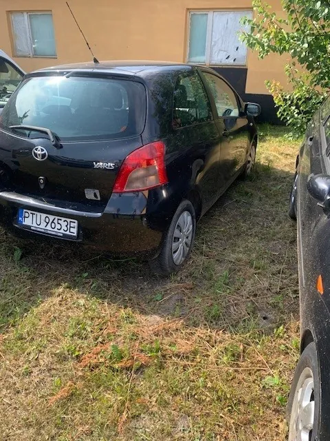 Czarny samochód marki Toyota Varis zaparkowany na trawiastym podłożu przed żółtym budynkiem z dwoma oknami. Widoczny jest tył i prawy bok pojazdu z polską tablicą rejestracyjną o numerze PTU 9653E. Po prawej stronie kadru widoczny jest fragment innego, ciemnego samochodu.