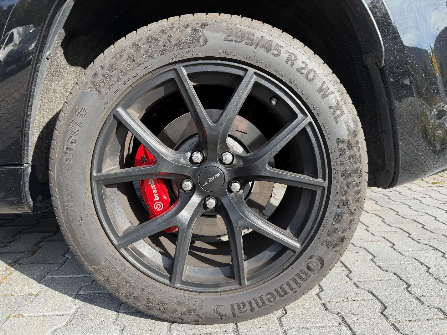 Zbliżenie na koło samochodu z czarną felgą o pięciu podwójnych ramionach i czerwonym zaciskiem hamulcowym z napisem 'brembo'. Opona marki Continental ma rozmiar 295/45 R 20 W XL. Na środku felgi znajduje się logo 'SRT'. Koło stoi na szarej kostce brukowej.