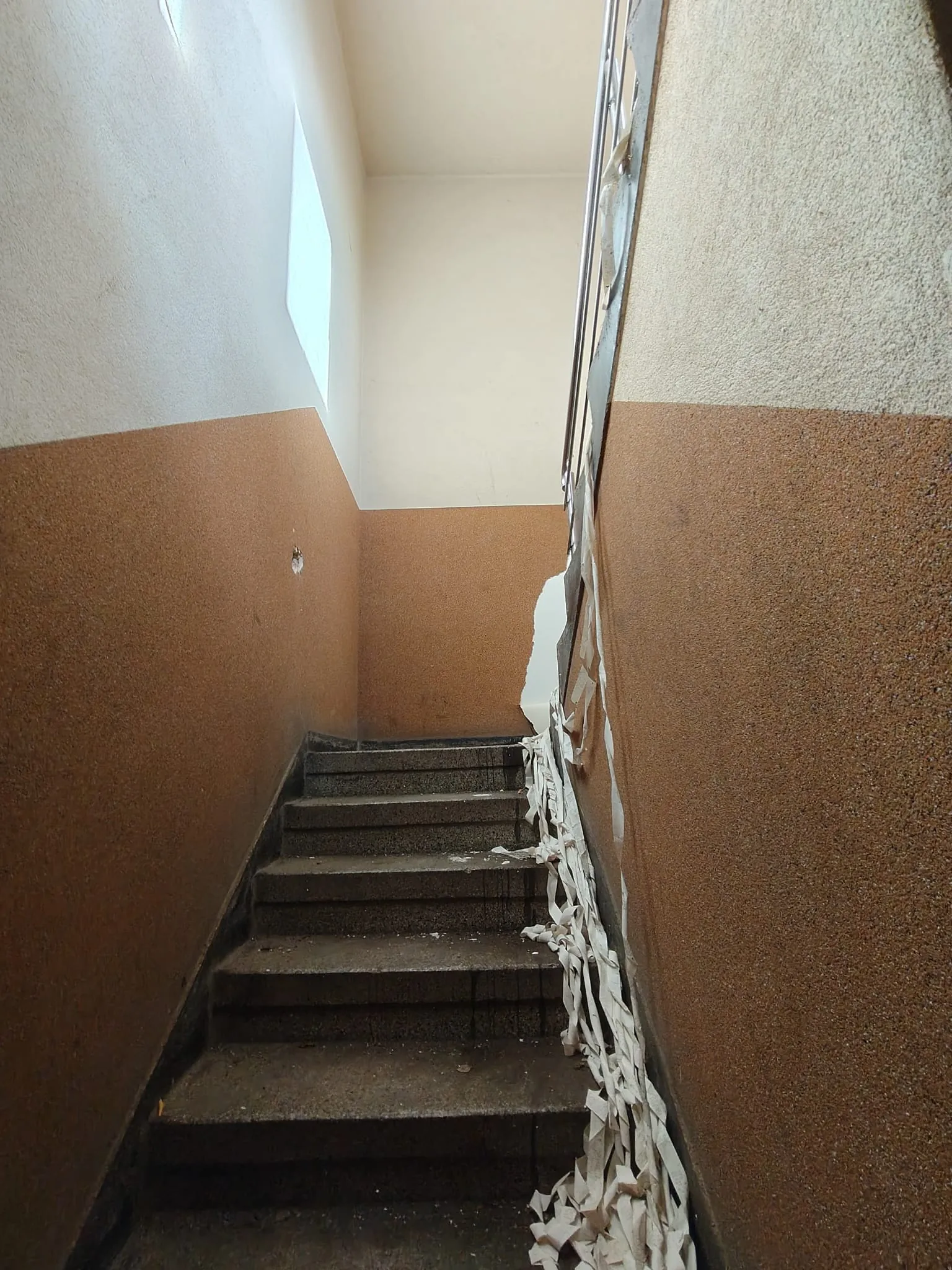 Schody wewnątrz budynku z ciemnoszarymi stopniami. Ściany są dwukolorowe: górna część jest biała, a dolna brązowa. Po prawej stronie ściany widoczne są resztki odrywającej się białej tapety lub tynku, które zwisają wzdłuż schodów. W górnej części lewej ściany znajduje się wąskie, pionowe okno wpuszczające światło.