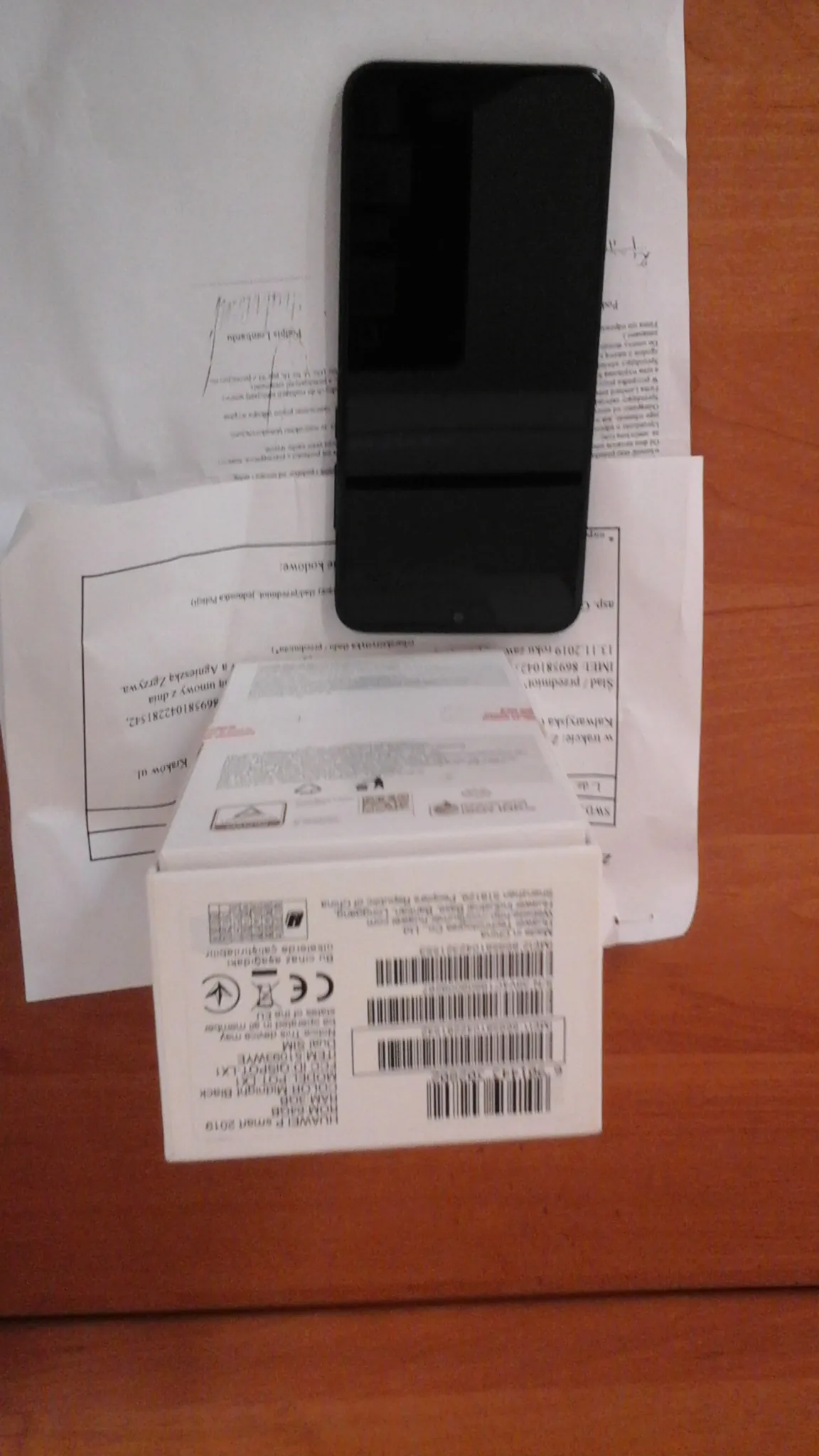 Czarny smartfon leżący na białych dokumentach z widocznym tekstem w języku polskim, w tym datą 13.11.2019 i fragmentami adresów. Obok smartfona znajduje się białe pudełko z kodami kreskowymi, oznaczeniem CE oraz napisem HUAWEI P smart 2019. Wszystko umieszczone na drewnianym blacie. Tekst na dokumentach i pudełku jest ułożony pionowo, co sugeruje nieprawidłową orientację obrazu.