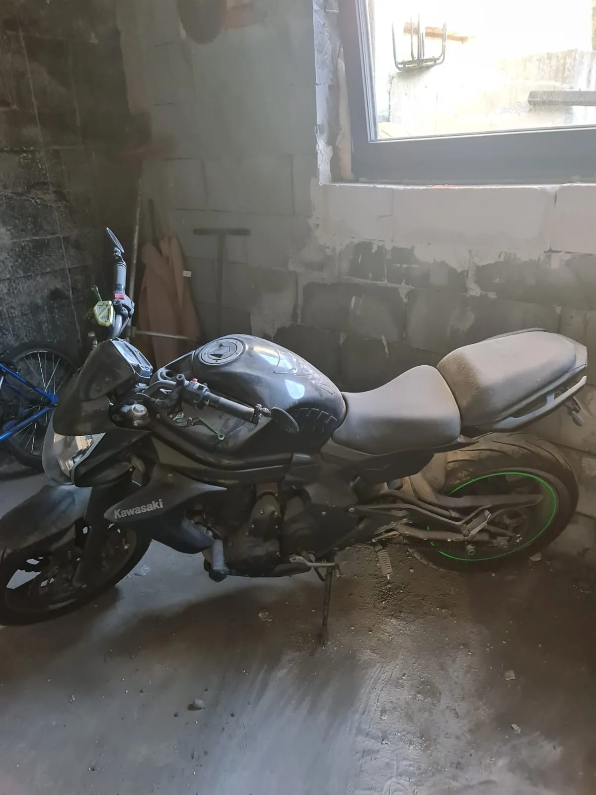 Czarny motocykl marki Kawasaki zaparkowany w pomieszczeniu z betonowymi ścianami. Widoczny zielony obrączek na tylnej oponie, czarne siedzenie i zbiornik paliwa. W tle okno, narzędzia oparte o ścianę oraz fragment niebieskiego roweru po lewej stronie. Podłoga jest betonowa, częściowo zabrudzona.