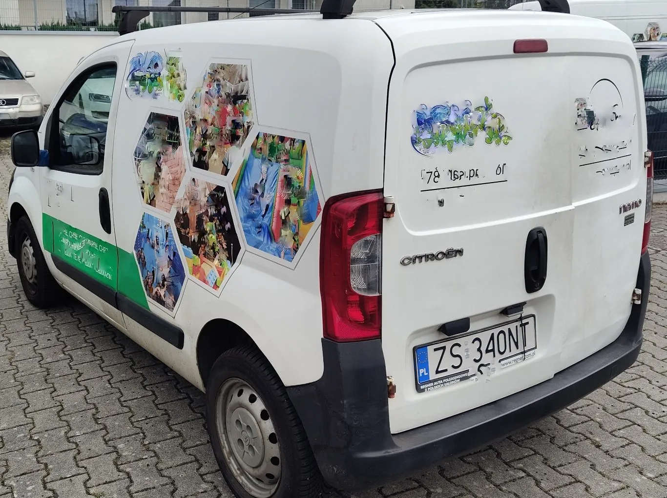 Biały samochód dostawczy marki Citroën zaparkowany na kostce brukowej. Na boku i tylnych drzwiach pojazdu znajdują się kolorowe naklejki w kształcie sześciokątów przedstawiające różne sceny oraz napisy. Na tylnych drzwiach widoczna jest tablica rejestracyjna o numerze ZS 340NT.