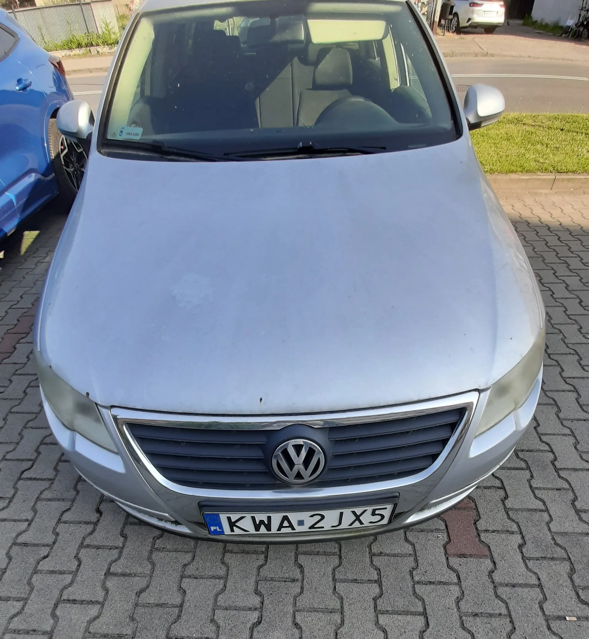 Srebrny samochód marki Volkswagen zaparkowany na kostce brukowej. Widok z przodu, z góry. Na masce widoczne są zmatowienia lakieru. Samochód posiada tablicę rejestracyjną o numerze KWA 2JX5.