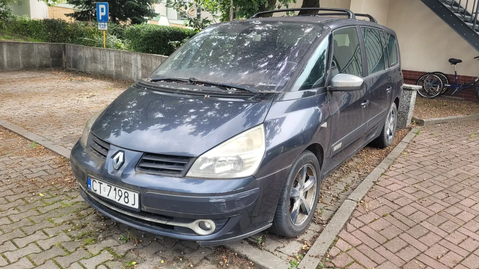 Ciemnoszary samochód marki Renault Scenic zaparkowany na kostce brukowej. Pojazd jest widoczny z przodu i z boku, posiada polską tablicę rejestracyjną o numerze CT 7198J. W tle widać zielone krzewy, drzewa oraz niebieski znak parkingowy z literą P.