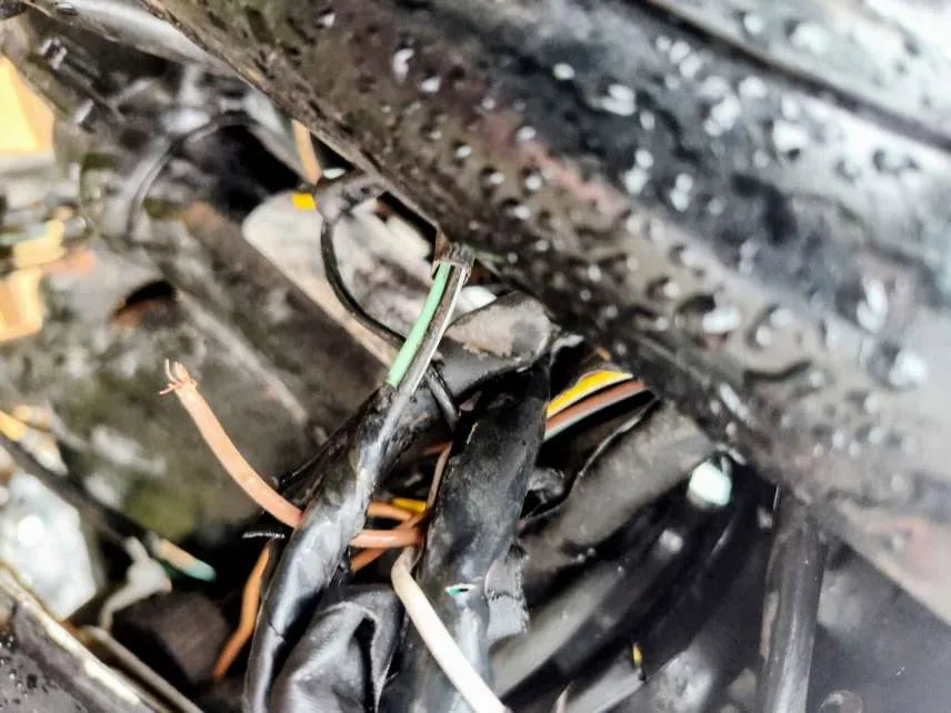Zbliżenie na uszkodzone przewody elektryczne o różnych kolorach: czarny, zielony, żółty, pomarańczowy i biały. Przewody są spalone, częściowo roztopione, z odsłoniętymi metalowymi żyłami. W tle widoczna metalowa powierzchnia pokryta kroplami wody lub kondensatu. Elementy są splątane, a izolacja niektórych przewodów jest uszkodzona.