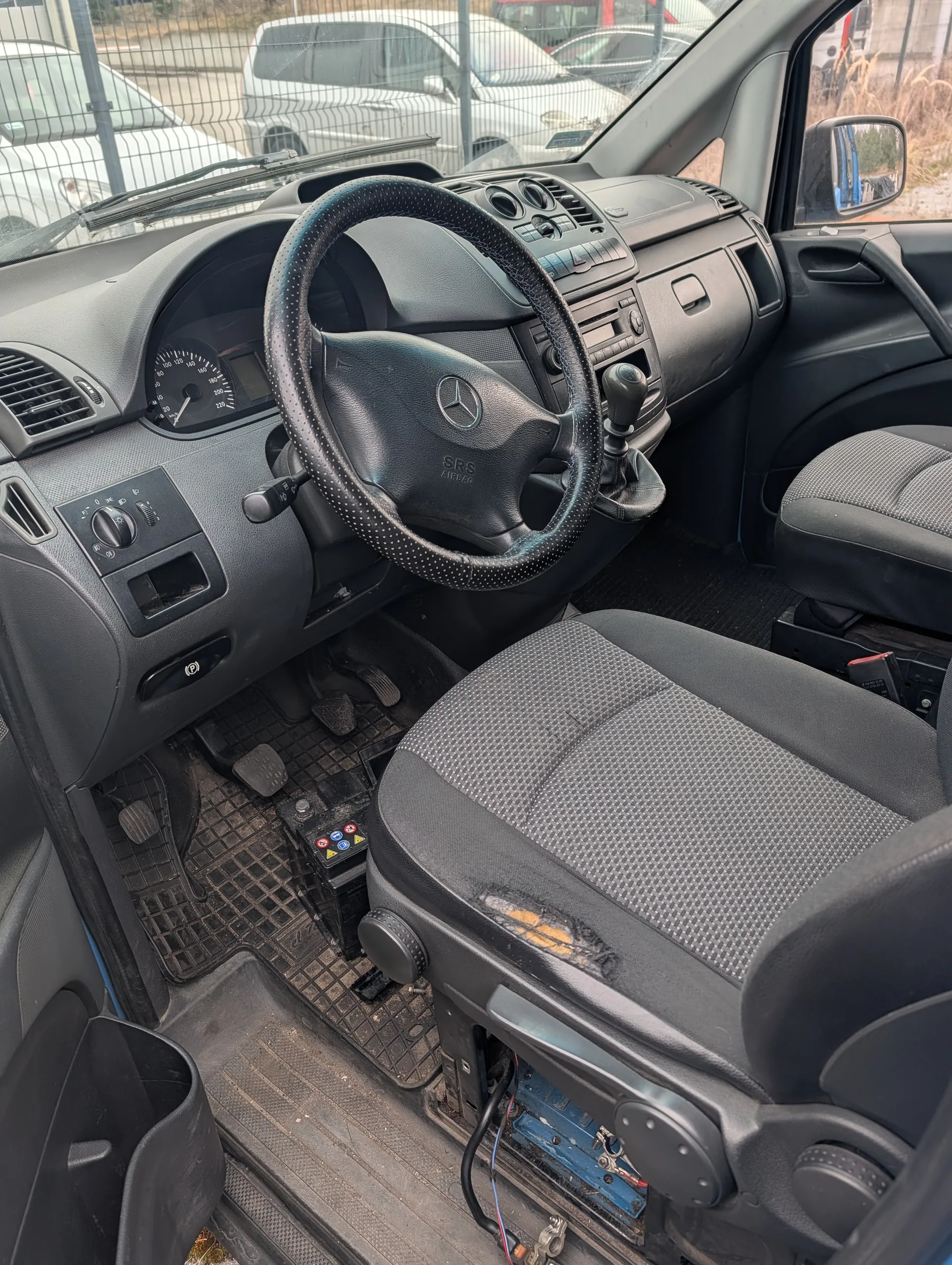 Wnętrze samochodu marki Mercedes, widok z pasażerskiego miejsca na kierowcę. Kierownica z logo Mercedes i napisem SRS AIRBAG, deska rozdzielcza z licznikiem prędkości, klimatyzacją i przyciskami. Siedzenia pasażerskie i kierowcy w szaro-czarnym kolorze, siedzenie kierowcy uszkodzone. Podłoga z czarnymi dywanikami, widoczne pedały i akumulator z kolorowymi przyciskami. Za przednią szybą widoczne zaparkowane samochody i ogrodzenie.