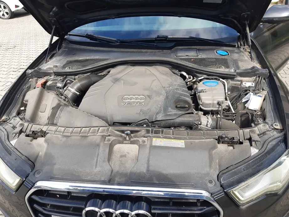 Widok z góry na otwartą maskę samochodu marki Audi, ukazującą komorę silnika. Centralnie znajduje się czarna osłona silnika z logo Audi i napisem „V6 TDI”. Wokół silnika widoczne są elementy układu chłodzenia, zbiornik płynu wycieraczek oraz przewody. Całość jest pokryta warstwą kurzu.