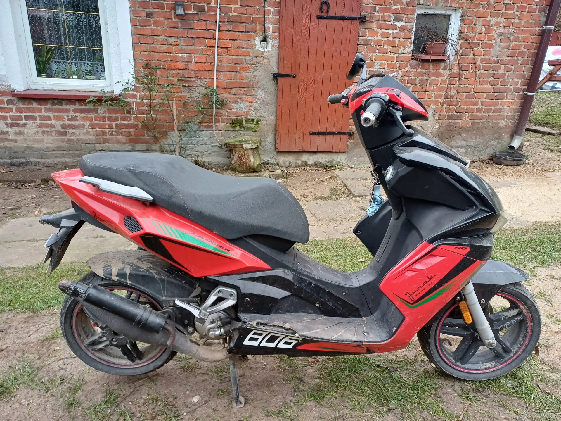 Rudno-czarny skuter z napisami "806", "50" i "Juno", zaparkowany na trawie i ziemi przed ceglanym budynkiem z białymi obramowaniami okien, czerwonymi drzwiami, roślinami w oknie i kamiennym blokiem. Maszyna zanieczyszczona, widoczne ślady brudu.