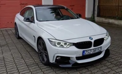 Biały samochód marki BMW coupe zaparkowany na kostce brukowej przed czerwonymi drzwiami garażowymi. Pojazd ma czarny dach, ciemne felgi i polską tablicę rejestracyjną o numerze ZSZ 38152.