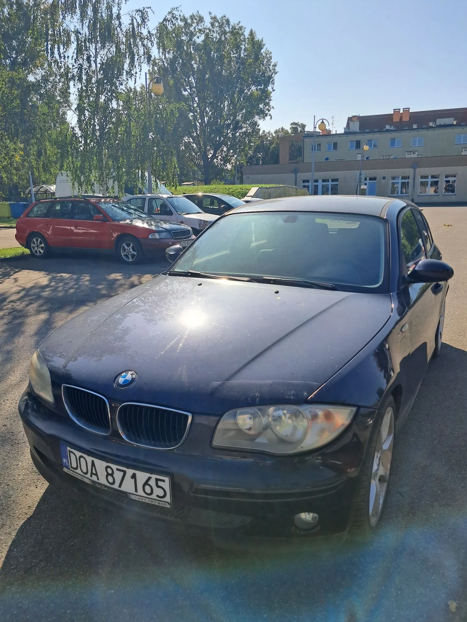 Czarny samochód marki BMW zaparkowany na zewnątrz. Na przednim zderzaku znajduje się tablica rejestracyjna o numerze DOA 87165. W tle widoczne są inne zaparkowane samochody, w tym czerwony i srebrny, zielone drzewa oraz jasny budynek mieszkalny lub biurowy pod bezchmurnym, niebieskim niebem.