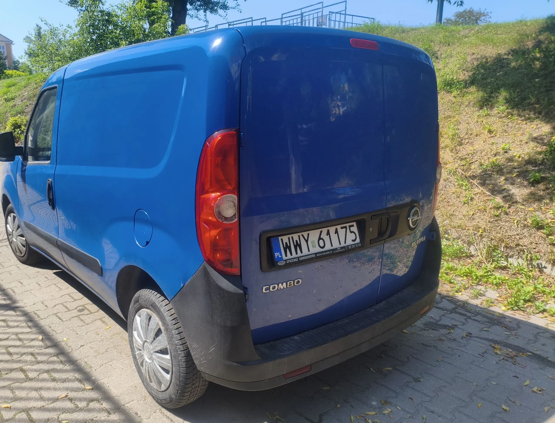 Niebieski samochód dostawczy marki Opel Combo zaparkowany na kostce brukowej. Widoczny jest tył pojazdu z tablicą rejestracyjną WWY 61175. Samochód stoi obok trawiastego nasypu w słoneczny dzień.