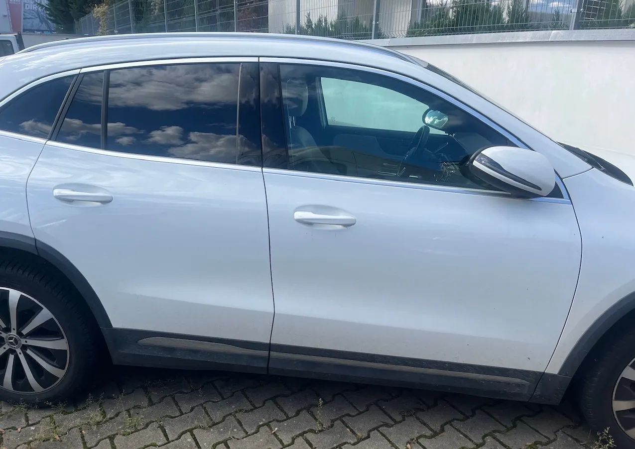 Boczny widok srebrnego samochodu typu SUV zaparkowanego na kostce brukowej. Widoczne są drzwi kierowcy i pasażera, lusterko boczne oraz felgi aluminiowe. W szybach odbija się niebo z chmurami.