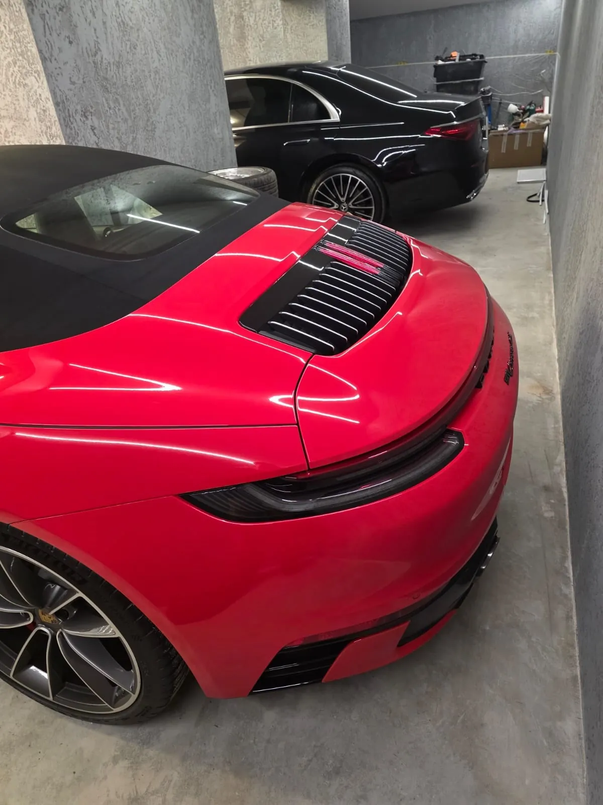 Czerwony samochód sportowy z widocznym tyłem, w tym reflektorem i klapą silnika z czarnymi listwami, zaparkowany w garażu. W tle czarny samochód osobowy, betonowe filary, podłoga oraz różne przedmioty warsztatowe. Na tylnej części czerwonego samochodu widoczny jest napis 'Taycan'.