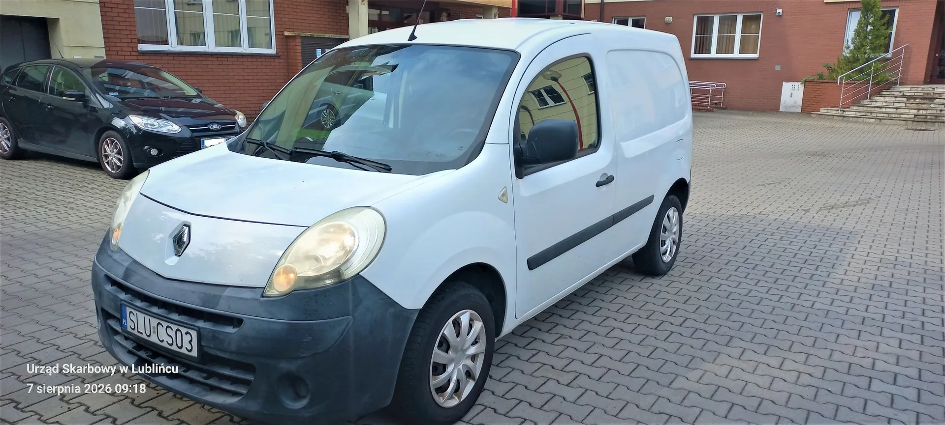Srebrny samochód dostawczy marki Renault zaparkowany na kostce brukowej przed budynkiem z czerwonej cegły. W tle widoczny jest czarny samochód osobowy oraz schody prowadzące do wejścia do budynku. W lewym dolnym rogu znajduje się napis: 'Urząd Skarbowy w Lublińcu 7 sierpnia 2026 09:18'. Tablica rejestracyjna samochodu to SLU CS03.