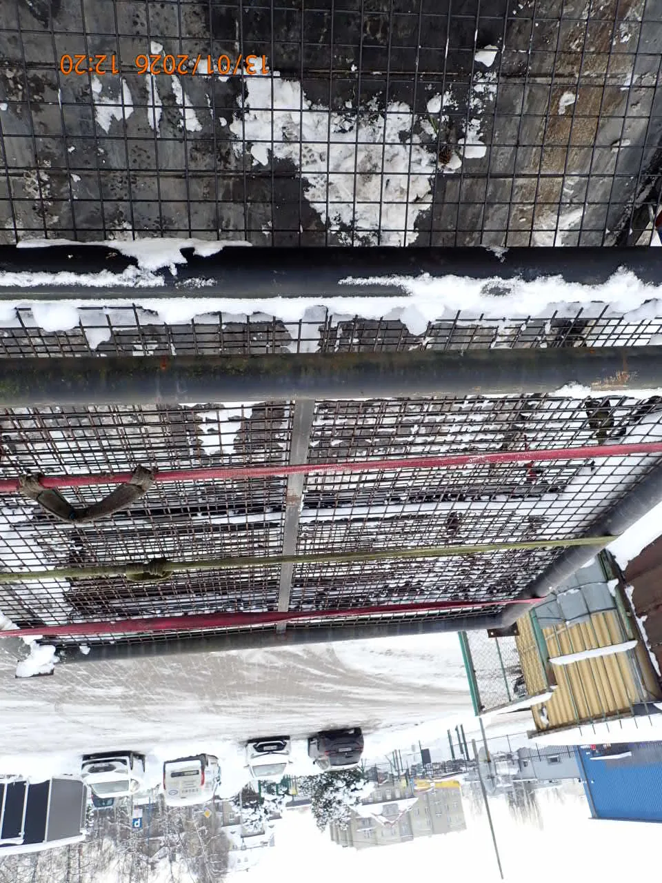 Czarna metalowa siatka z śniegiem na słupach i przęsłach, z czerwonymi i zielonymi paskami. Po lewej stronie widoczny pomarańczowy znaczek z datą i czasem: 13/01/2026 12:20. W tle zasypana śniegiem ulica z zaparkowanymi samochodami, budynkami i drzewami. Niebo jest pochmurne.