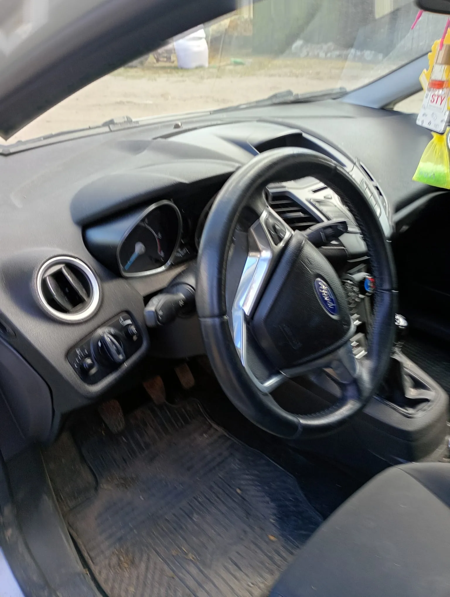 Wnętrze samochodu z widocznym kierownicą marki Ford, tablicą rozdzielczą z prędkościomierzem, klimatyzacją i przyciskami sterującymi. Na desce rozdzielczej widoczne są otwory wentylacyjne oraz pedały. Na podłodze czarne dywaniki z widocznymi zabrudzeniami. Przez przednią szybę widać zewnętrzny teren z suchą trawą i białym pojemnikiem. Na lusterku wstecznym wiszą żółty odświeżacz powietrza oraz tabliczka z napisem STY.