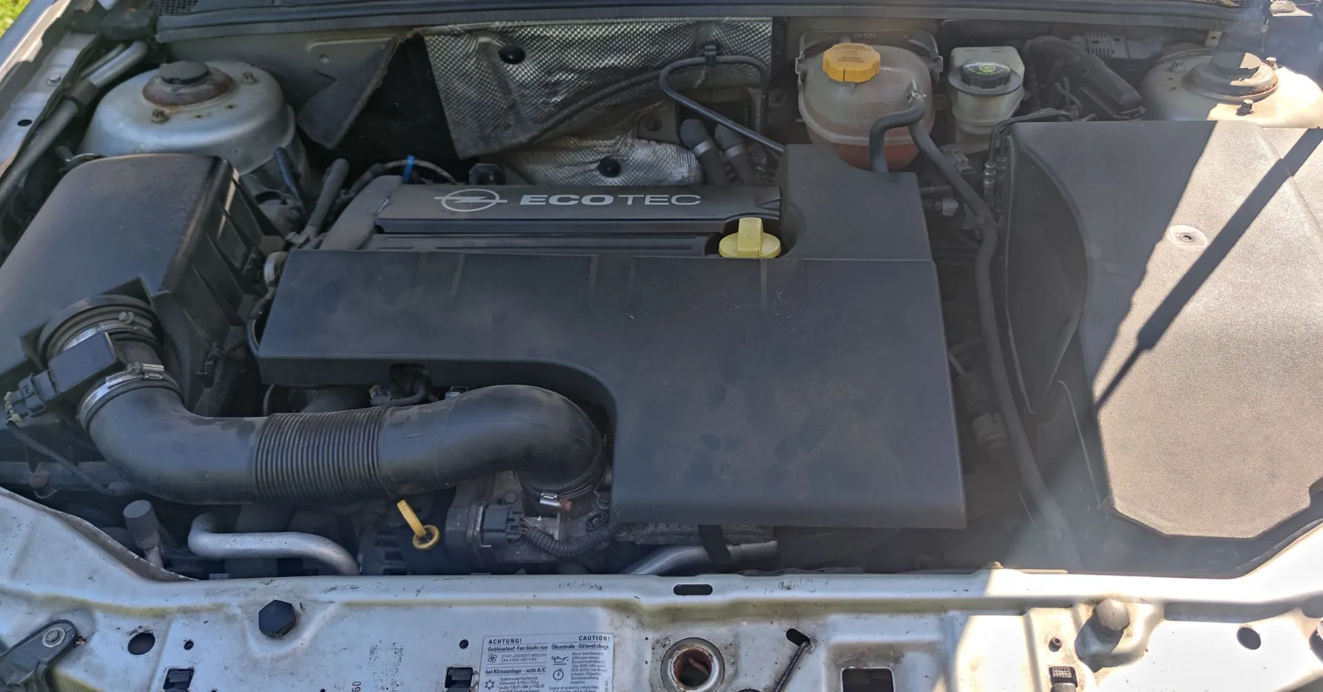 Widok z góry na komorę silnika samochodu. W centrum znajduje się czarna plastikowa osłona silnika z napisem 'ECOTEC' oraz logo marki Opel. Po prawej stronie widoczny jest zbiornik z płynem chłodniczym z żółtym korkiem, a po lewej gruby, czarny wąż dolotowy powietrza. Na dole zdjęcia widoczna jest srebrna karoseria z naklejką ostrzegawczą zawierającą tekst w języku niemieckim i angielskim.