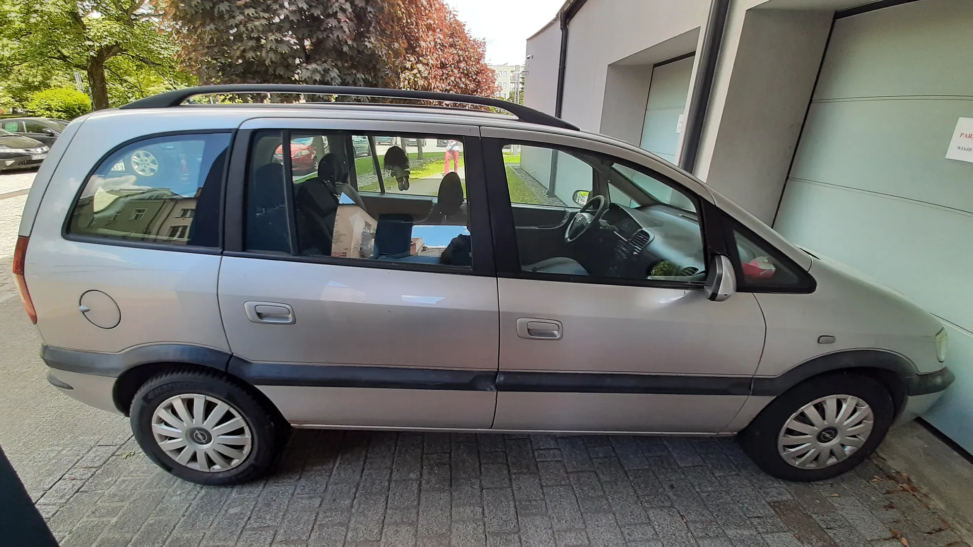 Srebrny minivan zaparkowany na brukowanej powierzchni obok jasnej budowli z częściowo widocznym napisem 'PARKING' na ścianie. Pojazd posiada czarny dachówkarz i srebrne felgi. Wewnątrz samochodu widoczne są przedmioty, w tym torba i ubrania. W tle drzewa z zielonymi i czerwonymi liśćmi oraz inne zaparkowane samochody. Budowla po prawej stronie ma szarą ścianę i białe drzwi garażowe.