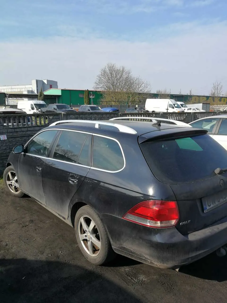 Ciemnoszary samochód osobowy typu kombi marki Volkswagen Golf zaparkowany na brudnej nawierzchni. Pojazd posiada białe dachowe pręty i tablicę rejestracyjną. W tle widoczne są inne samochody, kamienny mur z metalowym ogrodzeniem, zielony budynek z napisem OPALO oraz drzewa. Niebo jest częściowo zachmurzone.