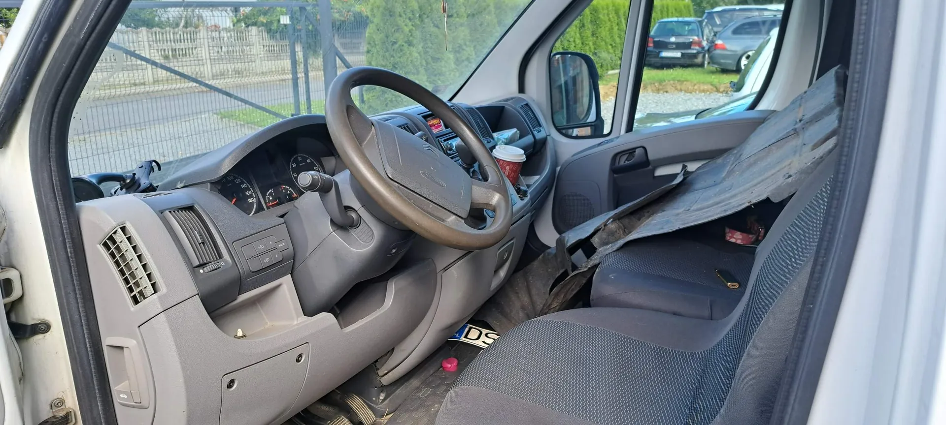 Wnętrze kabiny białego samochodu dostawczego widziane z perspektywy otwartych drzwi pasażera. Widoczna jest szara deska rozdzielcza, kierownica, panel instrumentów oraz dwa fotele. Na desce rozdzielczej znajduje się kubek z napojem. Pasażerski fotel jest częściowo przykryty ciemną, brudną płachtą materiału. Przez przednią szybę i okno widać zaparkowane samochody oraz ogrodzenie.