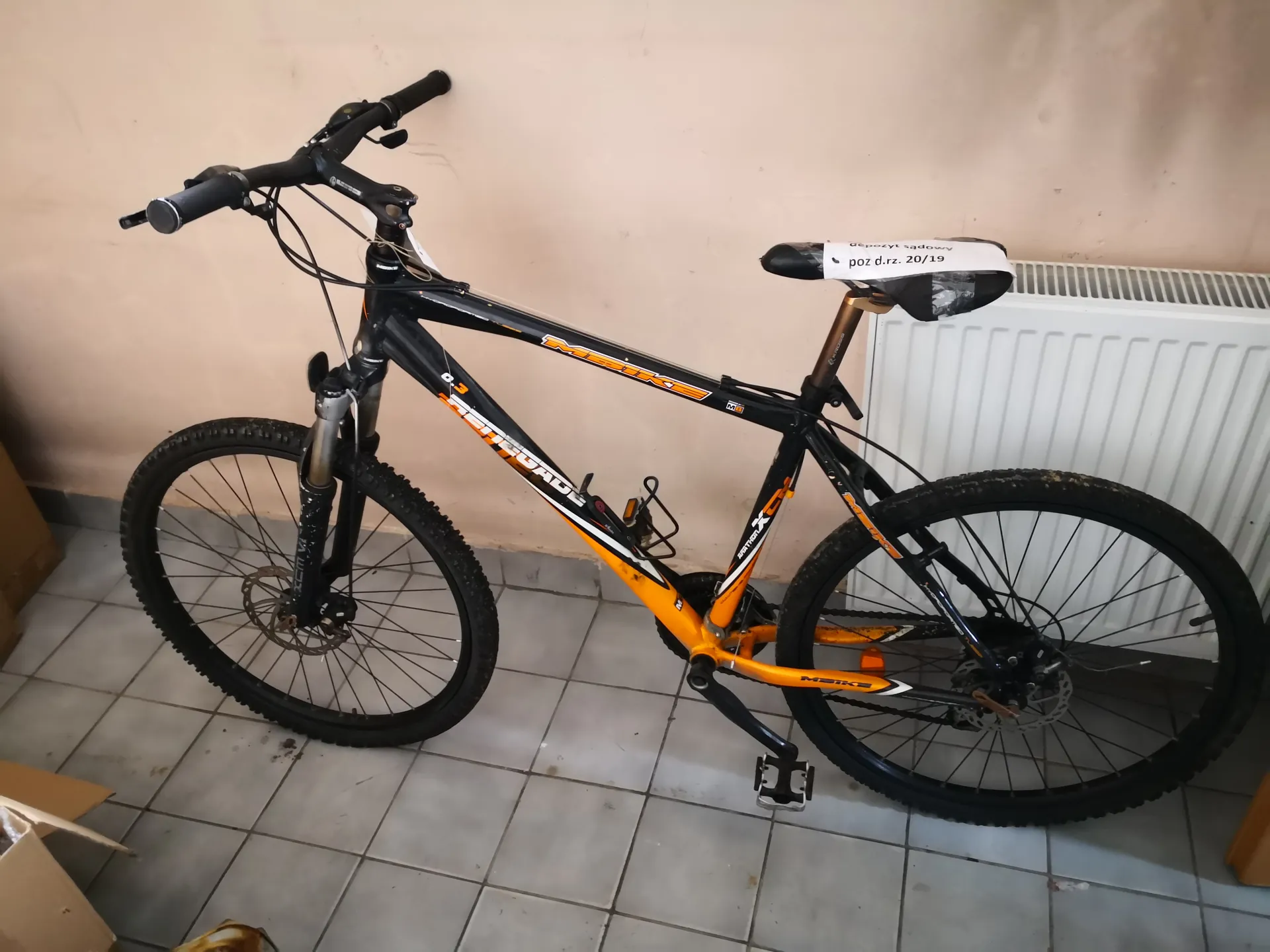 Rower górski marki Merida o czarno-pomarańczowej ramie, z widocznym napisem 'Merida' na ramie. Na siedzisku biała etykieta z napisem 'poz d.rz. 20/19'. Rower stoi na szarych płytkach podłogowych, przy beżowej ścianie z białym grzejnikiem po prawej stronie. W lewym dolnym rogu widoczne fragmenty kartonów.