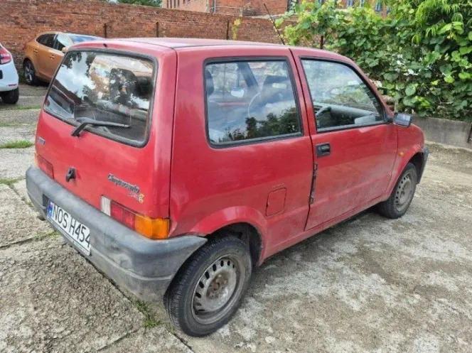 Czerwony, trzydrzwiowy samochód osobowy marki Fiat Cinquecento zaparkowany na betonowym podjeździe. Widoczny jest tył i prawy bok pojazdu. Samochód ma czarne plastikowe zderzaki i tablicę rejestracyjną o numerze NOS H454. W tle znajduje się ceglany mur oraz zielone krzewy.
