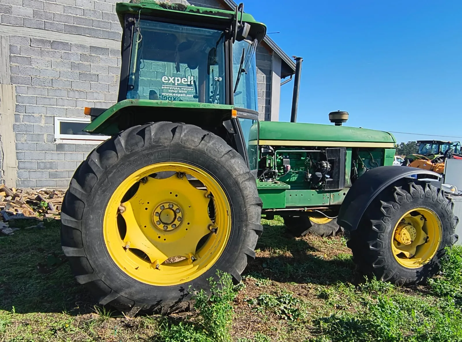 Zielony traktor z dużymi kołami z żółtymi felgami, zaparkowany na trawie przed szarym budynkiem z bloczków betonowych. Na przedniej szybie kabiny znajduje się biała naklejka z napisem 'expell' oraz adresem strony internetowej 'www.expell.net'. W tle widoczne jest bezchmurne, niebieskie niebo.