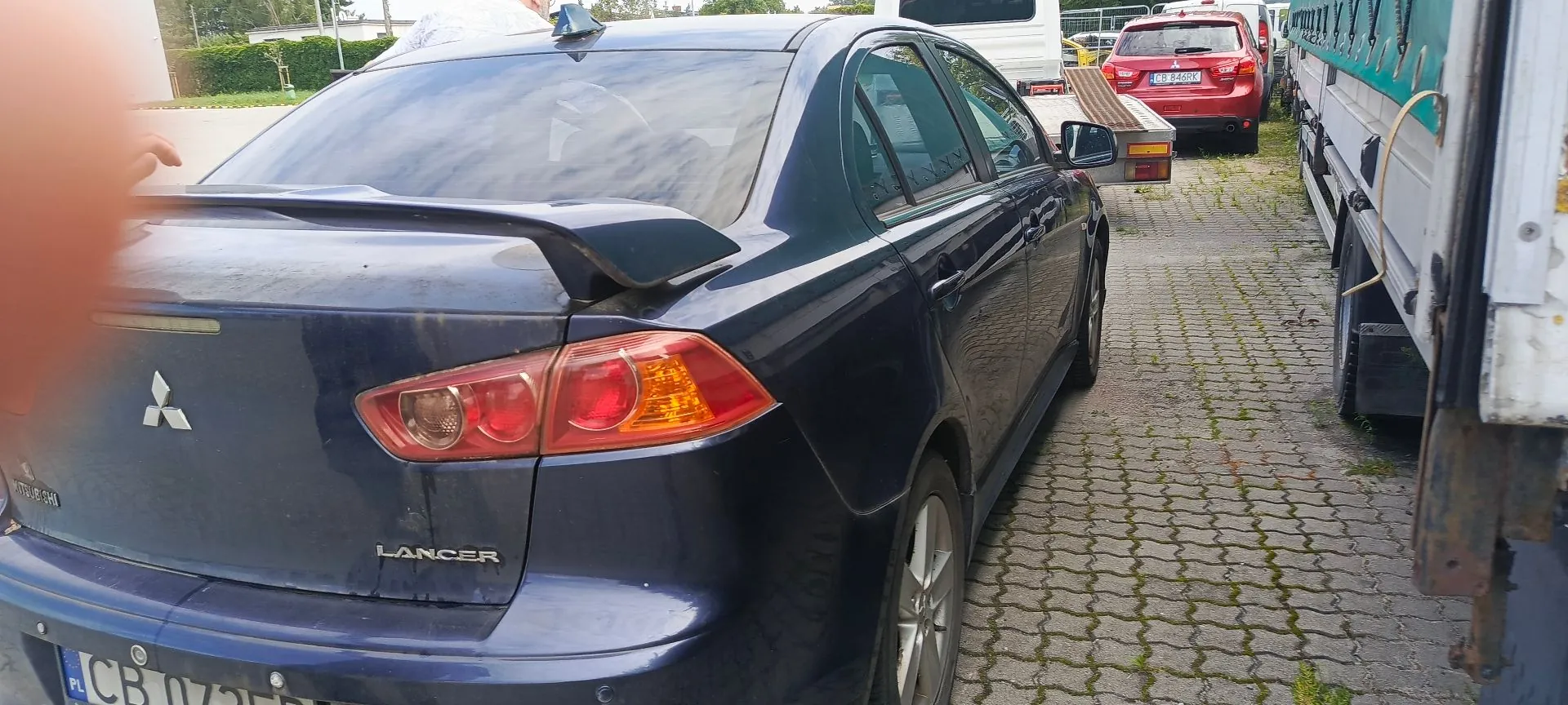 Tylna i boczna część ciemnoniebieskiego samochodu marki Mitsubishi Lancer zaparkowanego na kostce brukowej. Na klapie bagażnika widoczne jest logo Mitsubishi oraz napis LANCER. Samochód posiada spojler na klapie. W tle widoczny jest czerwony samochód oraz biały pojazd dostawczy.