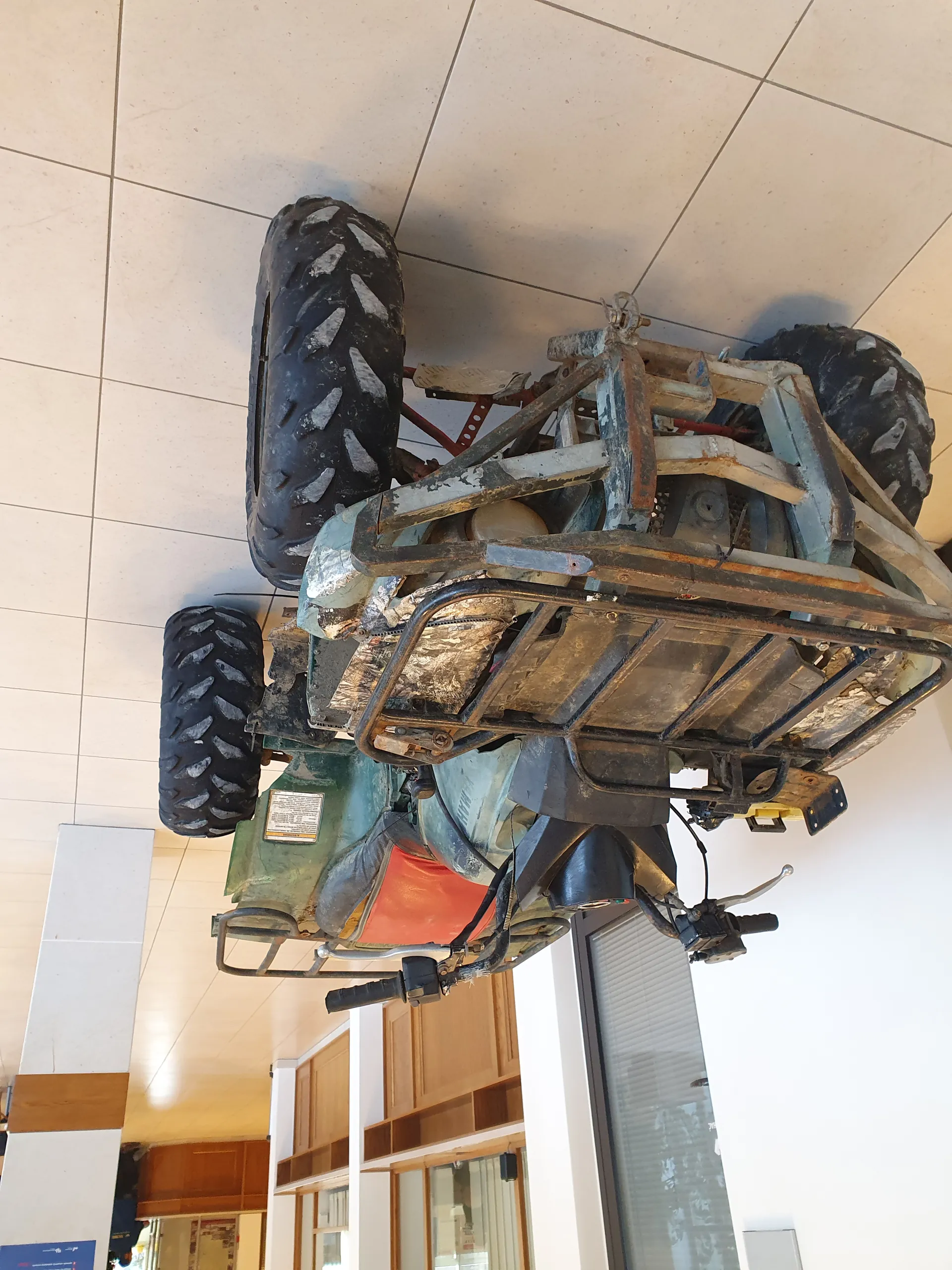 Zielony i czerwony quad z dużymi czarnymi oponami, umieszczony na boku na jasnej płytkowej podłodze. W tle widoczne są drewniane szafy, okna oraz biała ściana z niebieskim znakiem. Quad posiada metalową ramę, kierownicę i tabliczkę z informacjami na tylnej części.