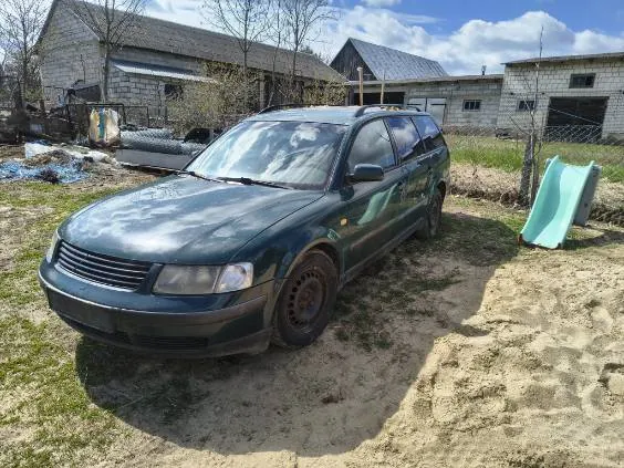 Zielony samochód typu kombi zaparkowany na piaskowatej i trawiastej powierzchni. W tle widoczne są budynki z cegły i dachówkami, w tym dom z dachem pokrytym blachą oraz struktura przypominająca garaż. Po prawej stronie samochodu znajduje się niebieska plastikowa zjeżdżalnia. Na ziemi leżą resztki śniegu, a na niebie widoczne są chmury. Drzewa bez liści rosną w tle.