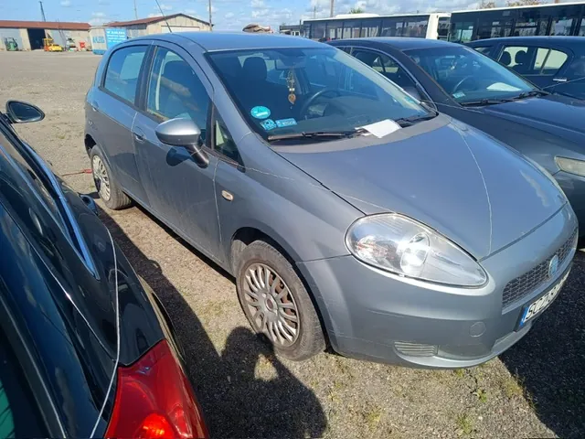 Srebrnoszary samochód osobowy marki Fiat Punto zaparkowany na żwirowym placu obok innych pojazdów. Auto jest widoczne z przodu i z boku. W tle znajdują się niskie budynki gospodarcze oraz biały autobus.