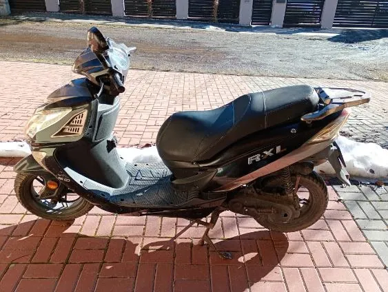 Skuter czarno-srebrny model RX1 zaparkowany na czerwonej chodnikowej kostce. W tle budynki z zamkniętymi żaluzjami i resztki śniegu przy krawężniku. Widoczny napis 'RX1' na boku skutera.