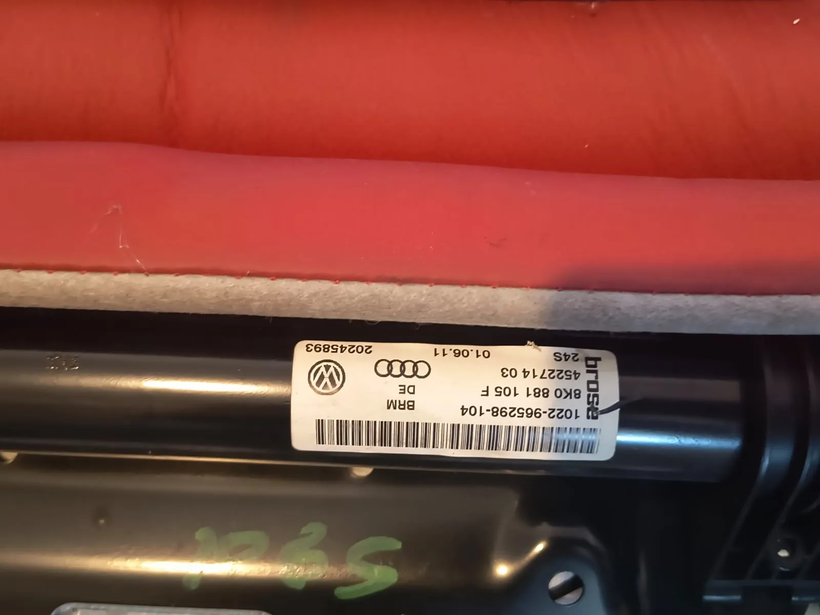 Zbliżenie na czarną, metalową część samochodową z naklejoną białą etykietą. Na etykietecie widoczne są logo Volkswagena i Audi oraz teksty: '1022-9652398-104', '8K0 881 105 F', '4522714 03', '24S', 'brose', 'BRIM DE', '01.06.11' oraz numer '20245893'. Obok części znajduje się czerwony element materiałowy.