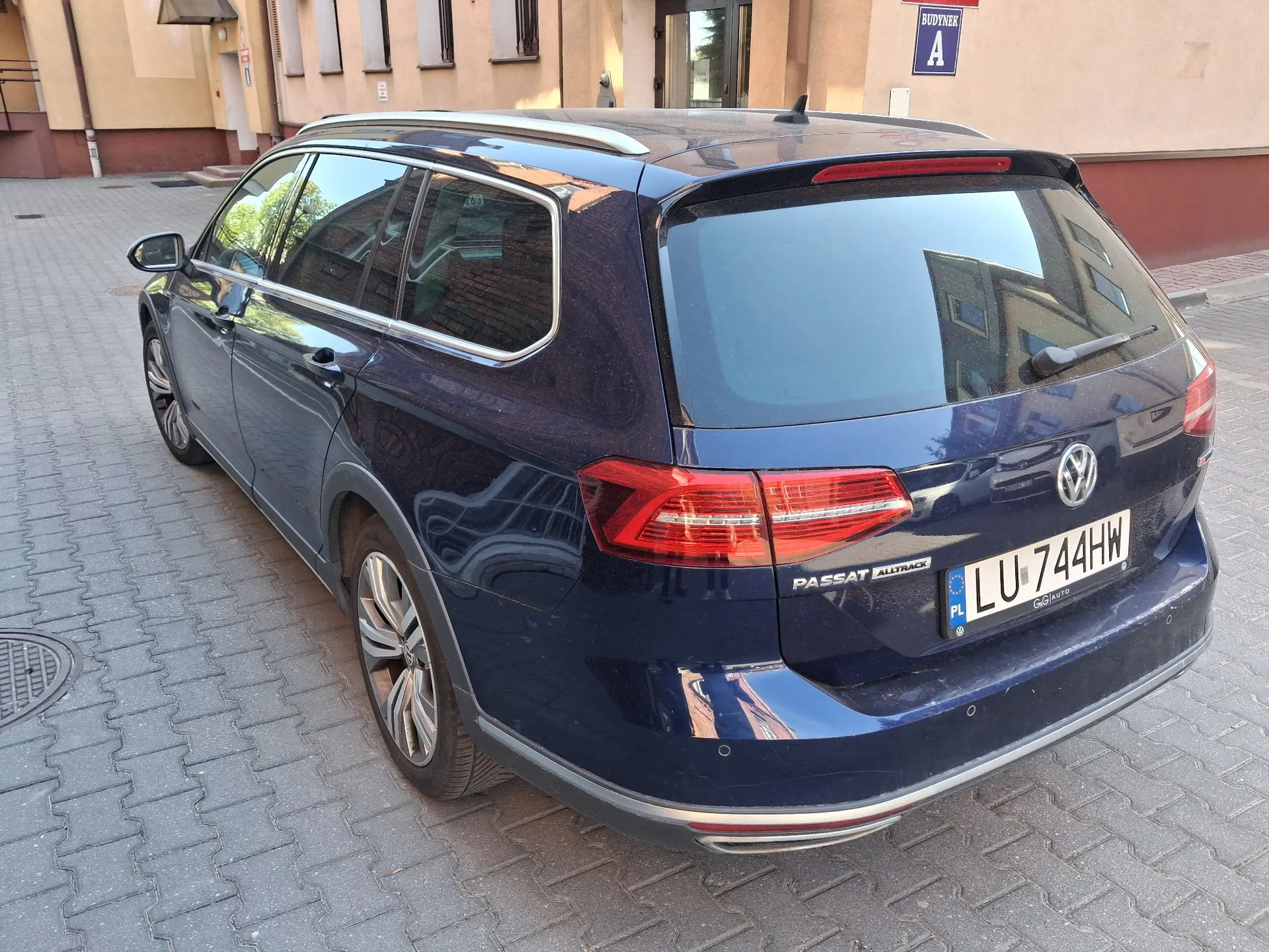 Granatowy samochód marki Volkswagen Passat Alltrack zaparkowany na kostce brukowej przed budynkiem. Widok z tyłu i z boku auta. Samochód ma polską tablicę rejestracyjną o numerze LU 744HW. W tle widoczna jest ściana budynku z niebieską tabliczką z napisem 'BUDYNEK A'.