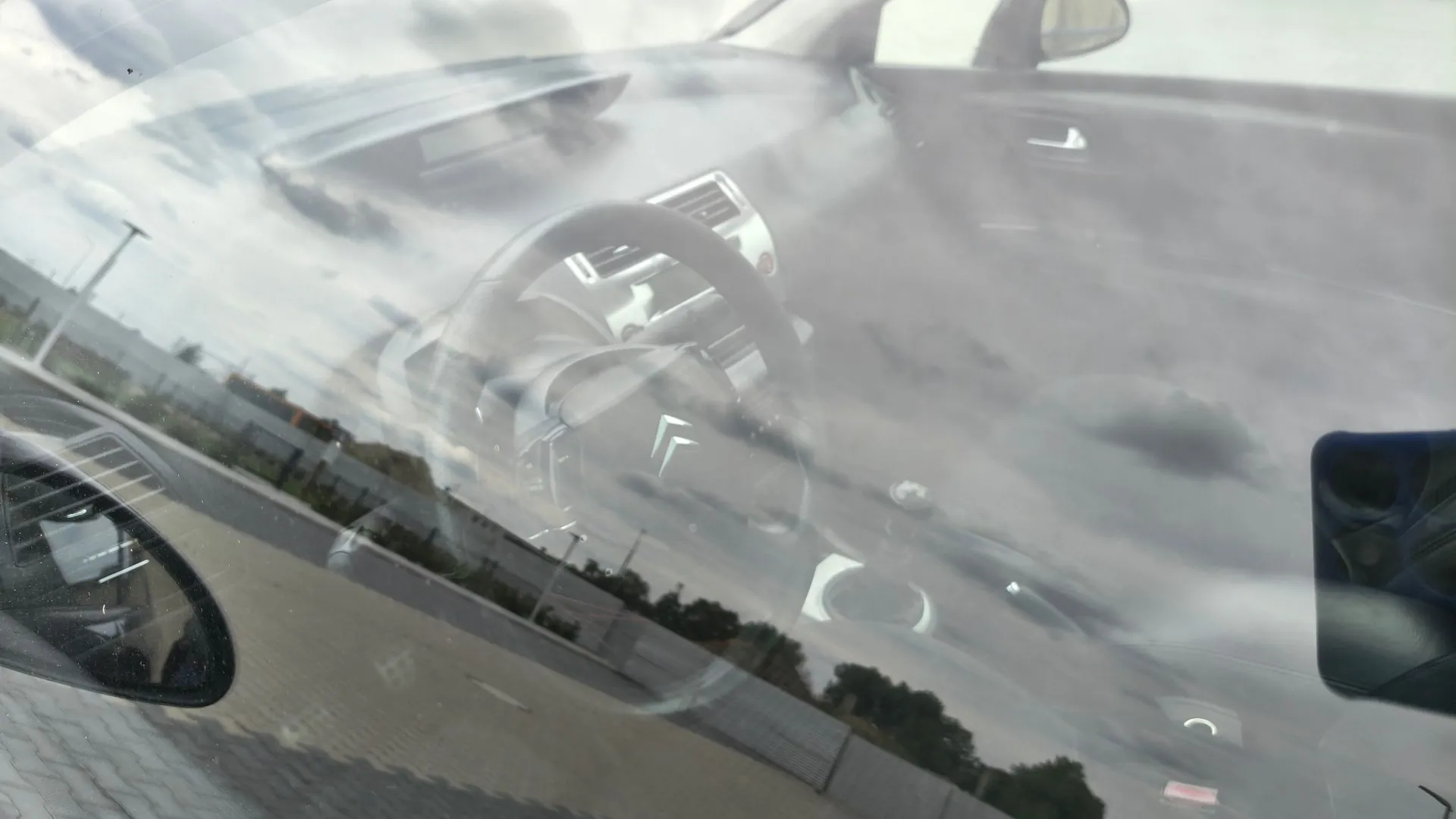 Zdjęcie wykonane przez szybę, na której widoczne są silne odblaski chmur i nieba. Przez szybę prześwituje wnętrze samochodu, w tym kierownica z logo marki Citroën oraz fragment deski rozdzielczej. W tle widać fragmenty ulicy i innych pojazdów.