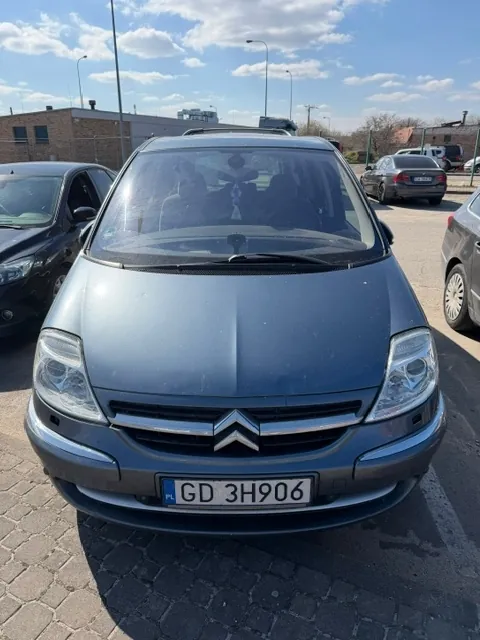 Zdjęcie przodu szarego samochodu marki Citroën zaparkowanego na kostce brukowej. Samochód ma polską tablicę rejestracyjną o numerze GD 3H906. Po lewej stronie widoczny jest fragment czarnego samochodu, a po prawej fragment srebrnego auta. W tle znajduje się budynek z cegły, latarnie uliczne oraz błękitne niebo z lekkimi chmurami.