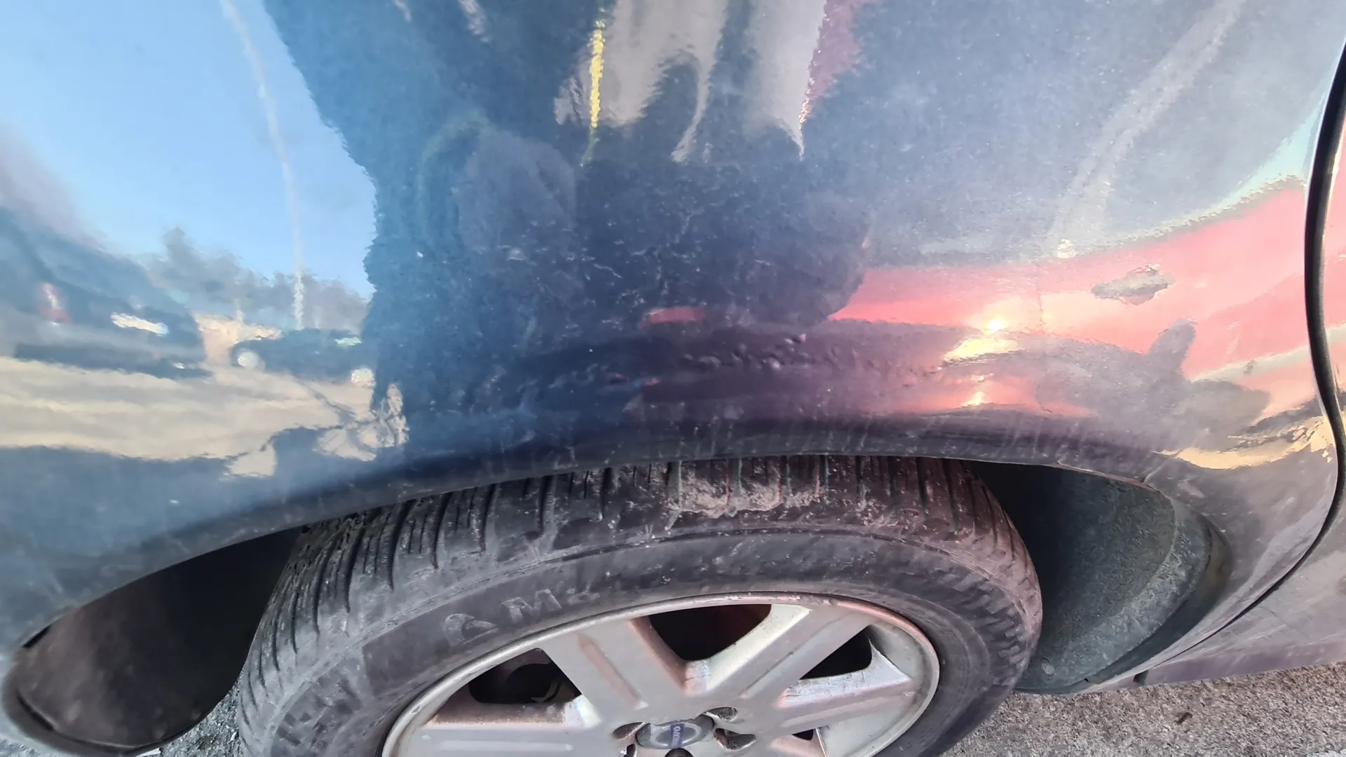 Zbliżenie na tylną część ciemnego samochodu z widocznym kołem. Opona jest zabrudzona, a felga ma srebrny kolor. Na karoserii odbijają się niebo i otoczenie. Podłoga z asfaltu.