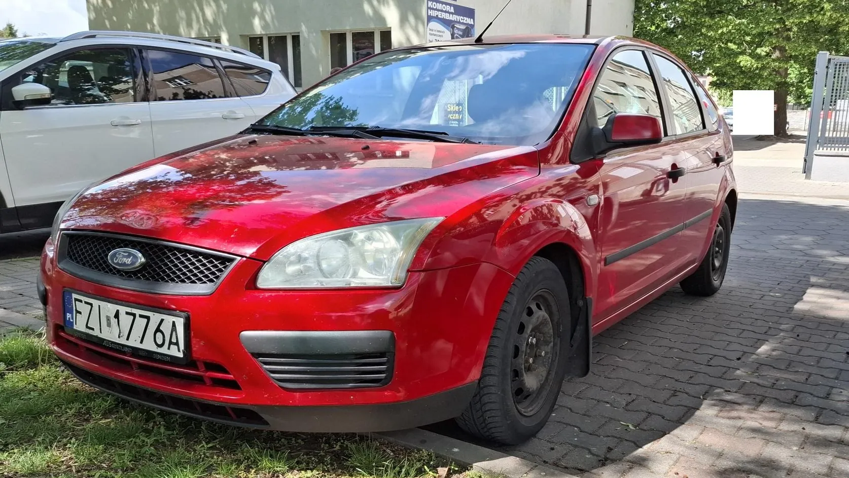 Czerwony samochód marki Ford zaparkowany na kostce brukowej obok białego samochodu. Widoczna jest przednia część i prawy bok auta. Samochód posiada polską tablicę rejestracyjną o numerze FZI 1776A. W tle znajduje się jasny budynek z szyldem 'KOMORA HIPERBARYCZNA'.