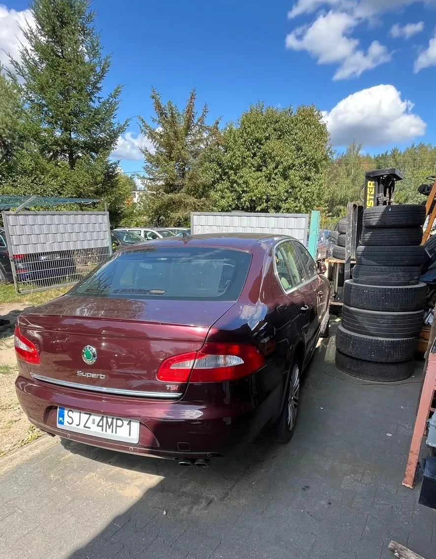 Tył bordowego samochodu marki Skoda Superb z tablicą rejestracyjną SJZ 4MP1. Auto zaparkowane jest na betonowym podłożu obok stosu czarnych opon. W tle widoczne są zielone drzewa, niebieskie niebo z białymi chmurami oraz metalowy ogrodzenie.