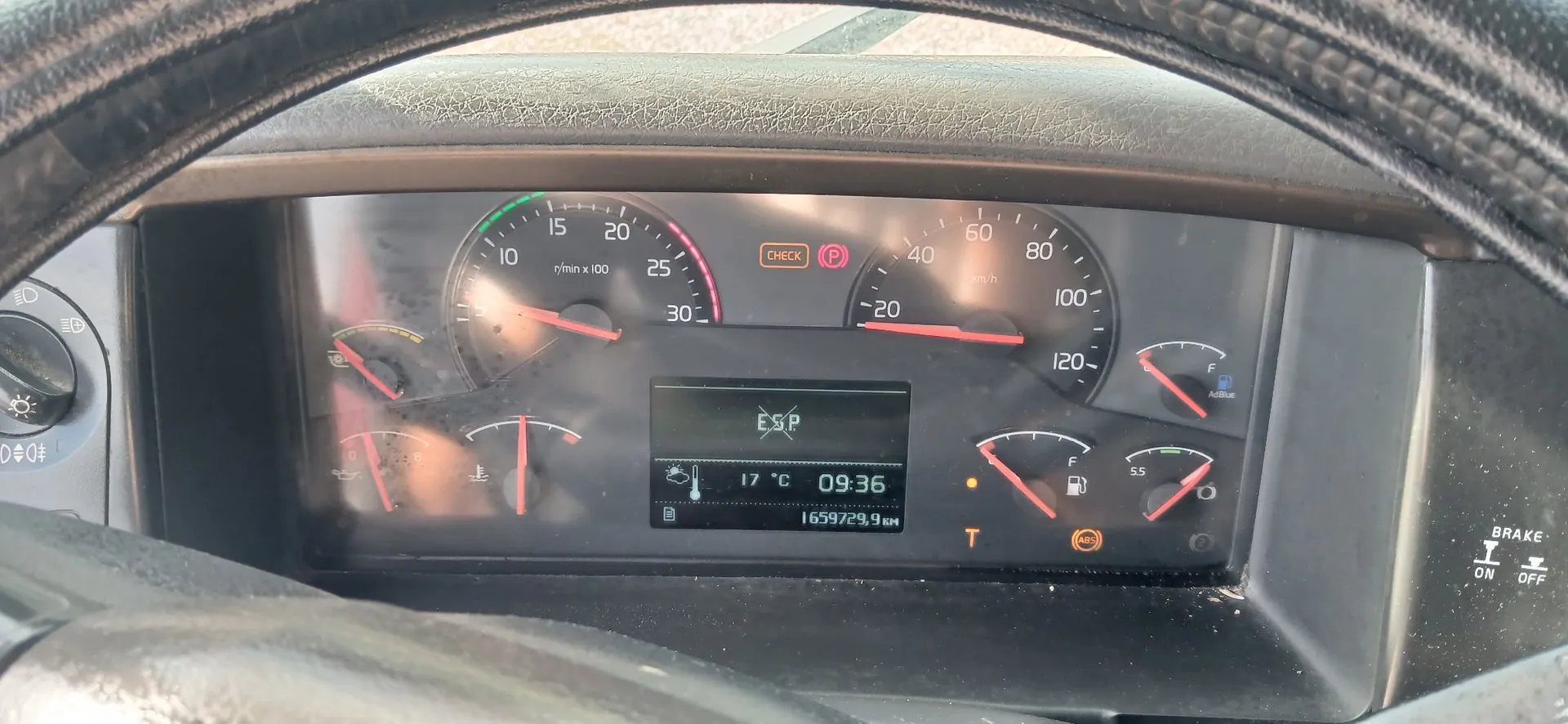 Widok z perspektywy kierowcy na deskę rozdzielczą ciężarówki. Widoczny jest licznik obrotów (do 30 x 100 r/min), prędkościomierz (do 120 km/h) oraz mniejsze wskaźniki paliwa, temperatury i ciśnienia. Na środku znajduje się cyfrowy wyświetlacz z napisem 'ESP', temperaturą '17 °C', godziną '09:36' oraz przebiegiem '1659729,9 km'. Zapalone są kontrolki 'CHECK' oraz 'P'. W kadrze widoczne są fragmenty kierownicy oraz przełączniki po prawej stronie z napisem 'BRAKE ON OFF'.