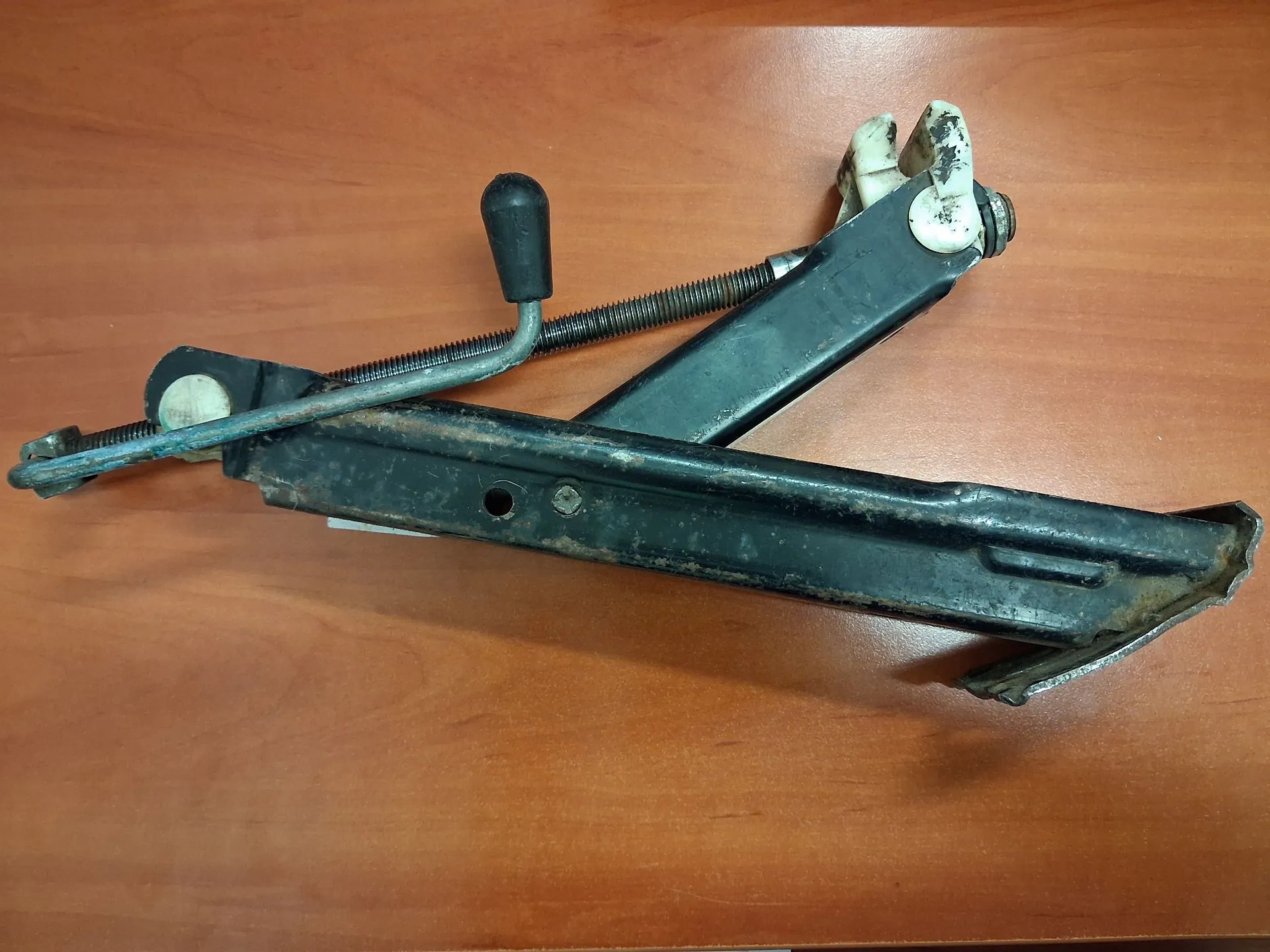 Metalowy mechanizm, prawdopodobnie część samochodowa, w kolorze czarnym z widocznymi śladami rdzy i zużycia. Element składa się z dwóch głównych ramion połączonych zawiasem, sprężyny oraz czarnej plastikowej rączki. Całość spoczywa na brązowej, drewnianej powierzchni.