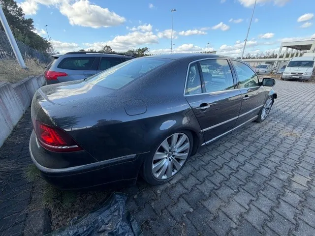 Czarny samochód osobowy typu sedan zaparkowany na brukowanej powierzchni. W tle widoczny szary SUV, biały van oraz betonowy barierka. Niebo z białymi chmurami, trawa przy barierce i słupy oświetleniowe. Samochód ma srebrne felgi i czerwone światła tylnego. Powierzchnia parkingu składa się z szarych, ząbkowanych płyt.