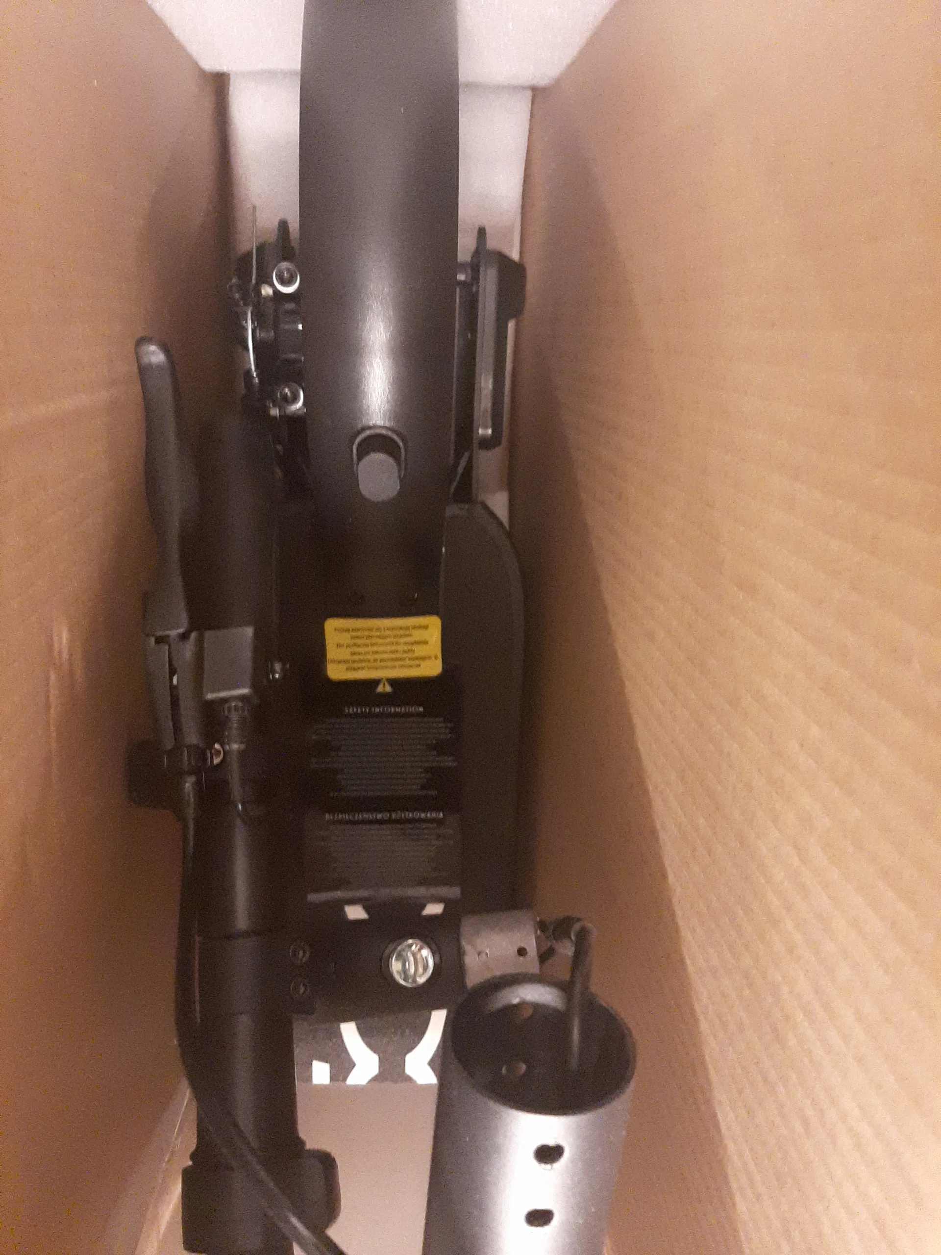 Część przodu czarnego elektrycznego skutera umieszczonego w brązowym kartonie. Widoczne są czarna rama, przednie koło, kierownica z reflektorem oraz czarna tabliczka z białym symbolem i żółtą ostrzegawczą etykietą. Tło stanowi brązowy karton z białą pianką u dołu.