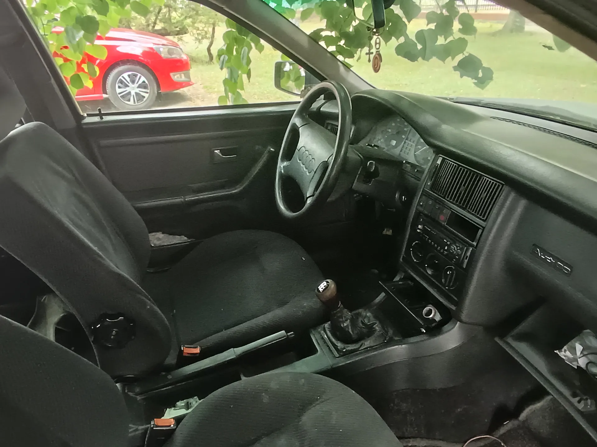 Wnętrze samochodu marki Audi z widocznym kierownicą z logo Audi, czarnymi siedzeniami, dźwignią zmiany biegów i deską rozdzielczą z radioodtwarzaczem. Przez przednią szybę i okno pasażera widoczny czerwony samochód zaparkowany na trawie oraz zielone liście drzew. Na lusterku wstecznym wiszą ozdoby, w tym krzyżyk. W prawym dolnym rogu deski rozdzielczej widoczny napis "Audi 80".