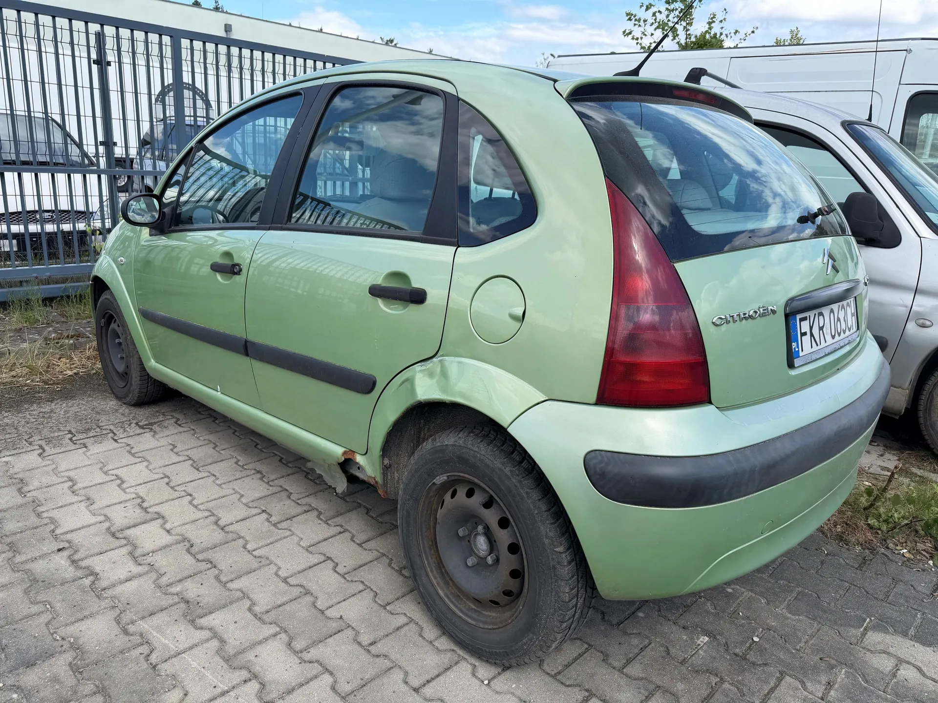 Zielony mały samochód osobowy marki Citroën z widocznym uszkodzeniem na tylnej prawej stronie nadwozia. Na tylnym zderzaku umieszczona tablica rejestracyjna z numerem FKR 063CH. Samochód zaparkowany na brukowanej powierzchni, w tle metalowe ogrodzenie, inne pojazdy oraz częściowo zachmurzone niebo. Widoczne są również czarne elementy dekoracyjne na bokach nadwozia i czerwone światła tylne.
