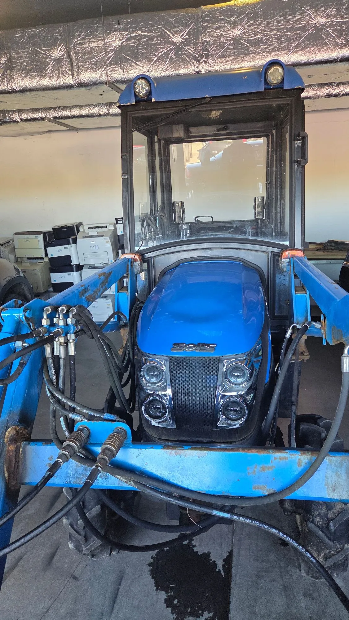 Niebieski traktor z przednim ładowaczem, zaparkowany w garażu lub magazynie. Maszyna posiada zamkniętą kabinę z dużymi szybami oraz widoczny napis 'SOIS' na przedniej masce. Z przodu widoczne są cztery reflektory oraz systemy hydrauliczne z przewodami. W tle znajdują się białe kartony i elementy konstrukcyjne pomieszczenia.