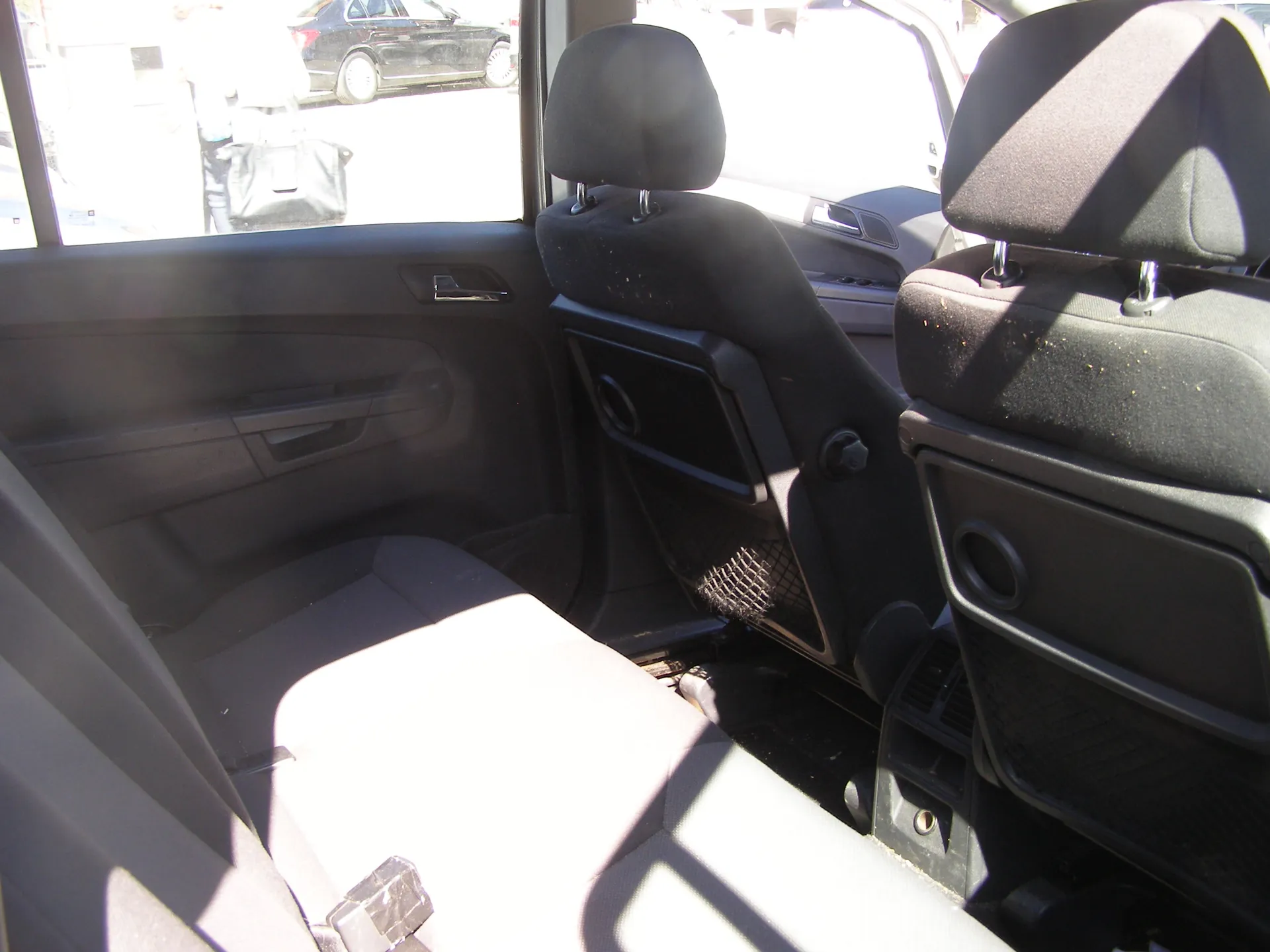 Wnętrze samochodu widziane z tyłu, przednie siedzenia z czarnymi zagłówkami i siateczkami na oparciach. Widoczne są fragmenty drzwi z uchwytem i oknem, przez które widać zewnętrzny parking z czarnym samochodem. Siedzenia mają ślady brudu, a wnętrze jest oświetlone słońcem rzucającym cienie.