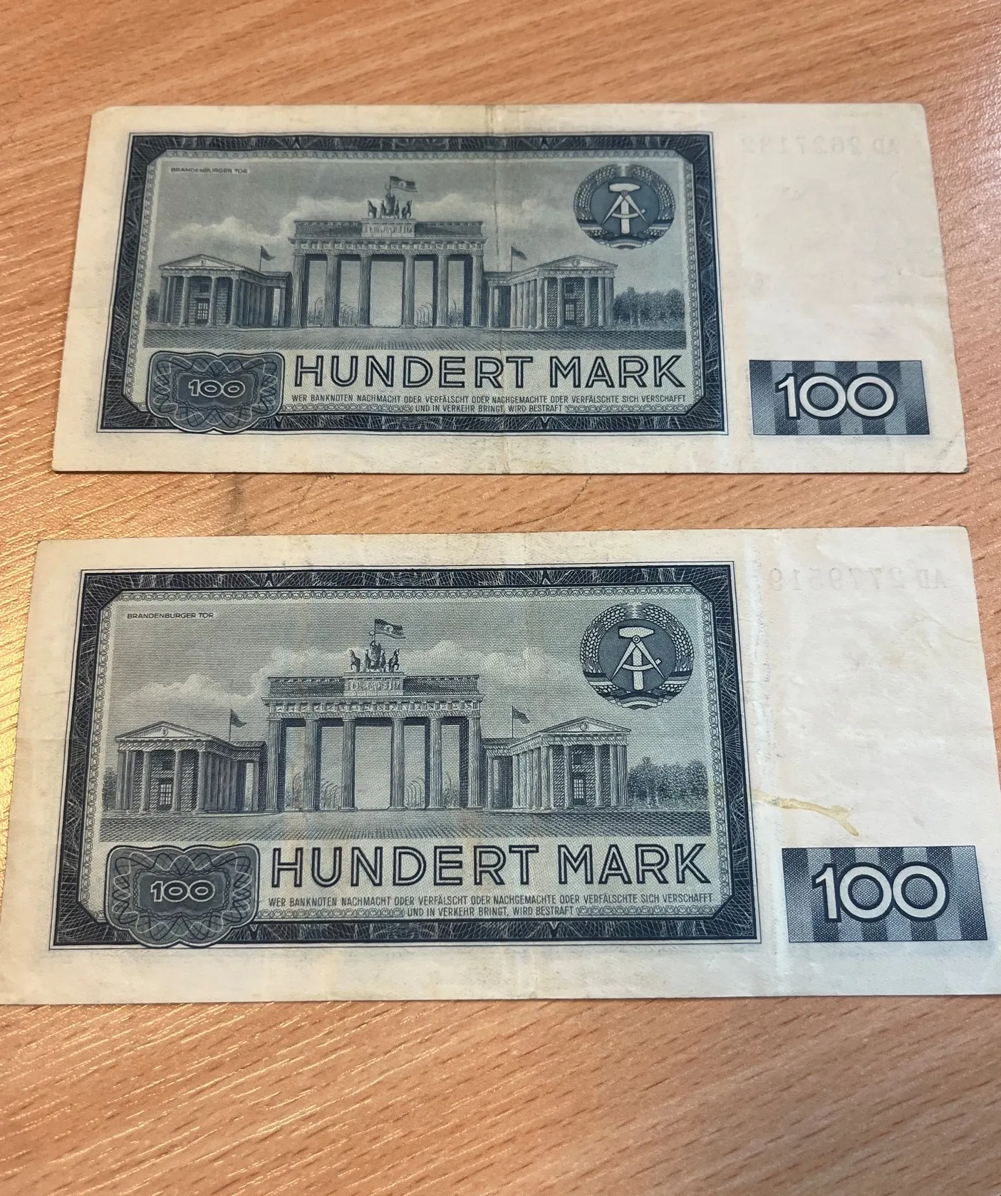 Dwa banknoty o nominale 100 marek (Hundert Mark) z Niemieckiej Republiki Demokratycznej, leżące jeden nad drugim na drewnianym blacie. Na banknotach widnieje wizerunek Bramy Brandenburskiej oraz godło NRD. W centralnej części znajduje się napis 'HUNDERT MARK', a w rogach liczba '100'.